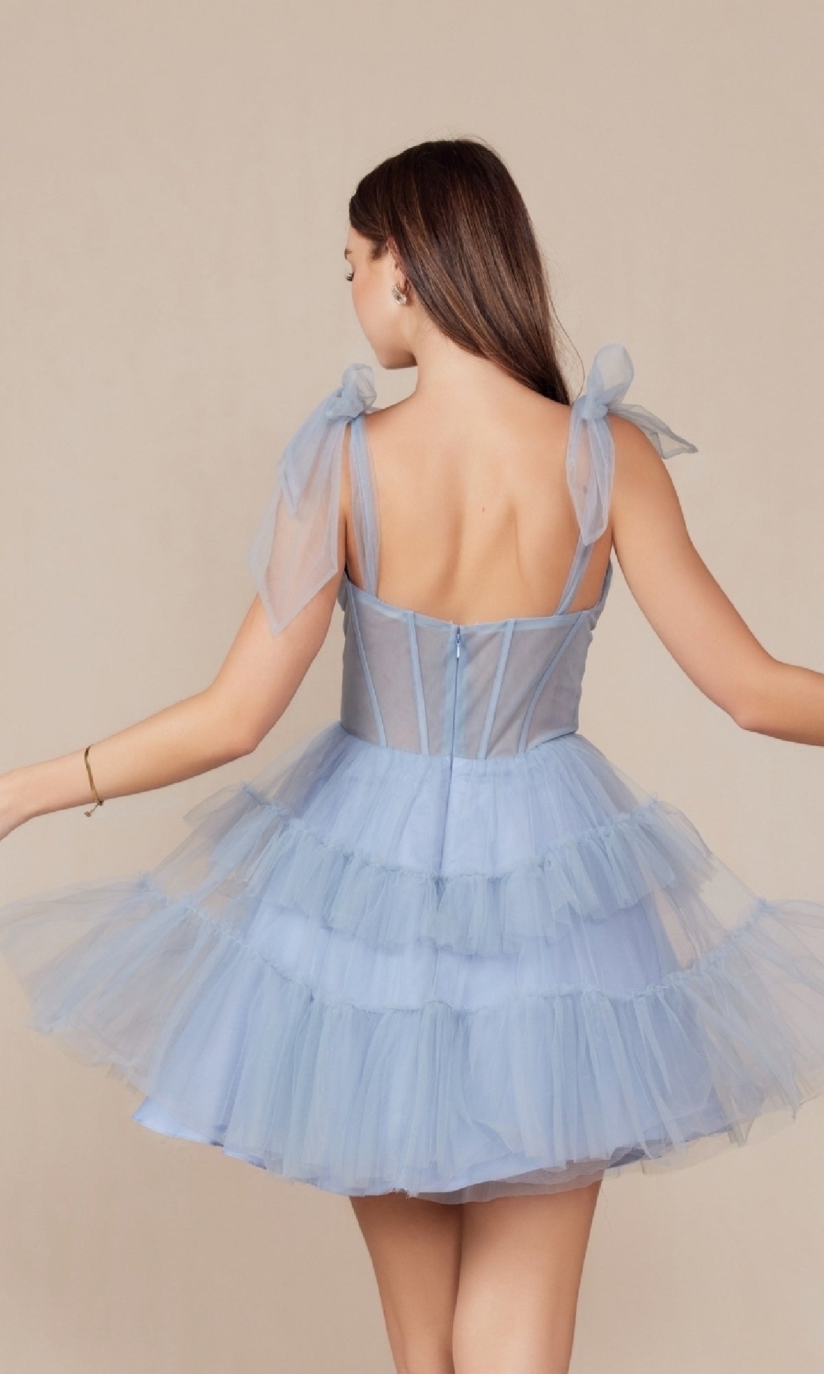 A woman faces away, wearing the Shoulder-Tie Short Ruffled A-Line Hoco Dress Y858. This light blue dress features shoulder bows, a sheer corset bodice, and a tiered flared skirt for an elegant party look.
